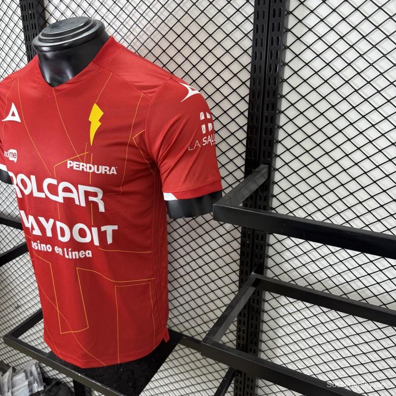 قميص اللاعب رقم 25/26 للنادي نيكاكسا (خارج أرضه) باللون الأحمر مع نقوش هندسية ذهبية على شكل صواعق